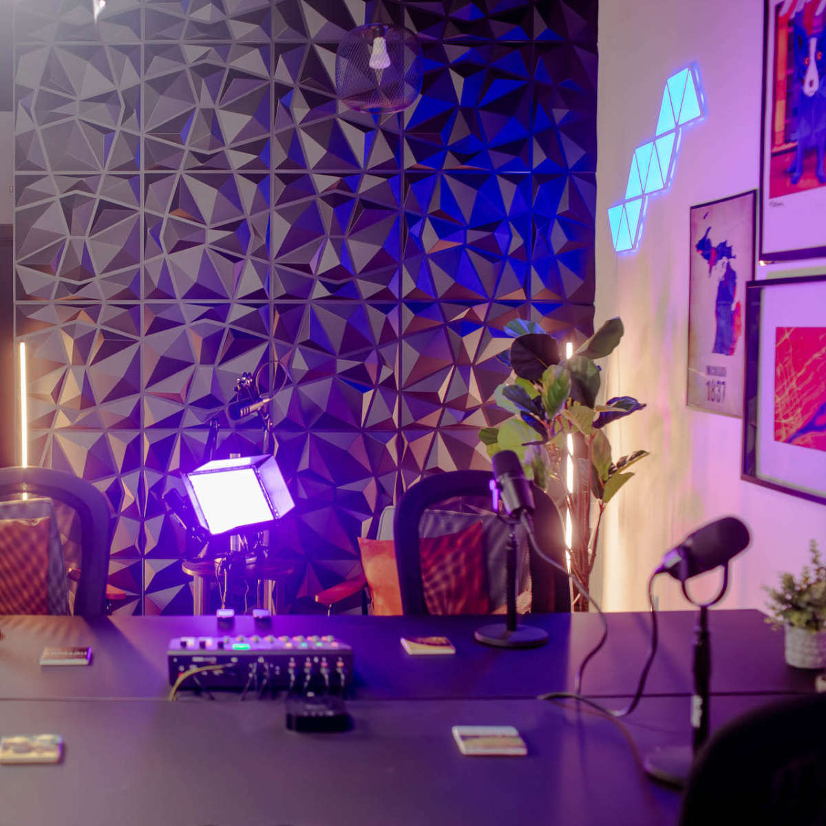 A modern podcast studio setup featuring a table with microphones, a sound mixer, and a geometric patterned wall illuminated by colorful lights.