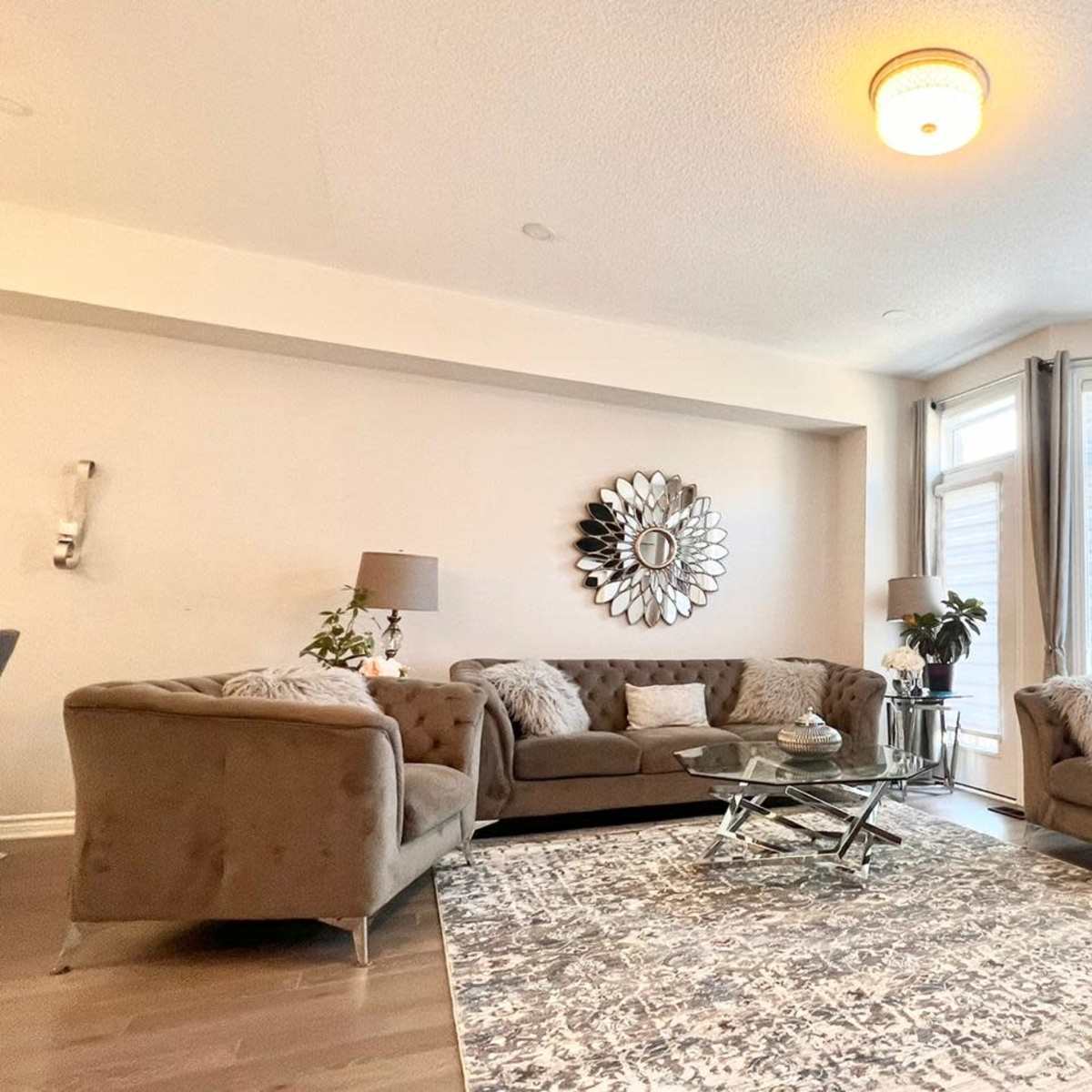 A modern living room features a sectional sofa, a glass coffee table, decorative pillows, and large windows with sheer curtains.
