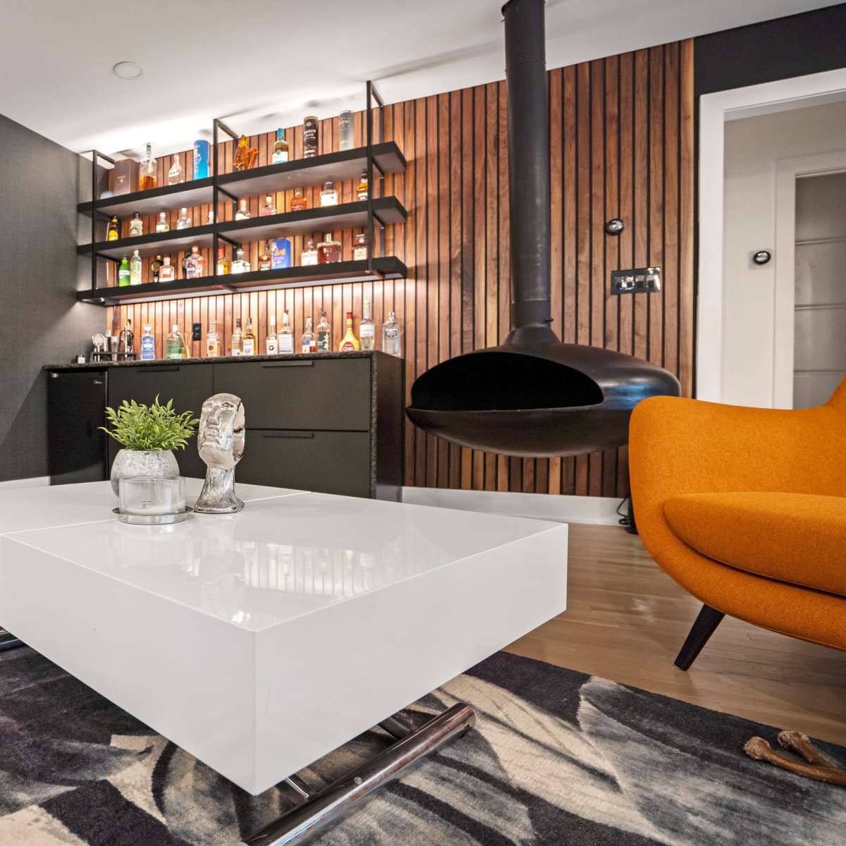A modern living room with a gray wall, a white coffee table, an orange chair, and a black fireplace against a wooden accent wall displaying a shelf of bottles.