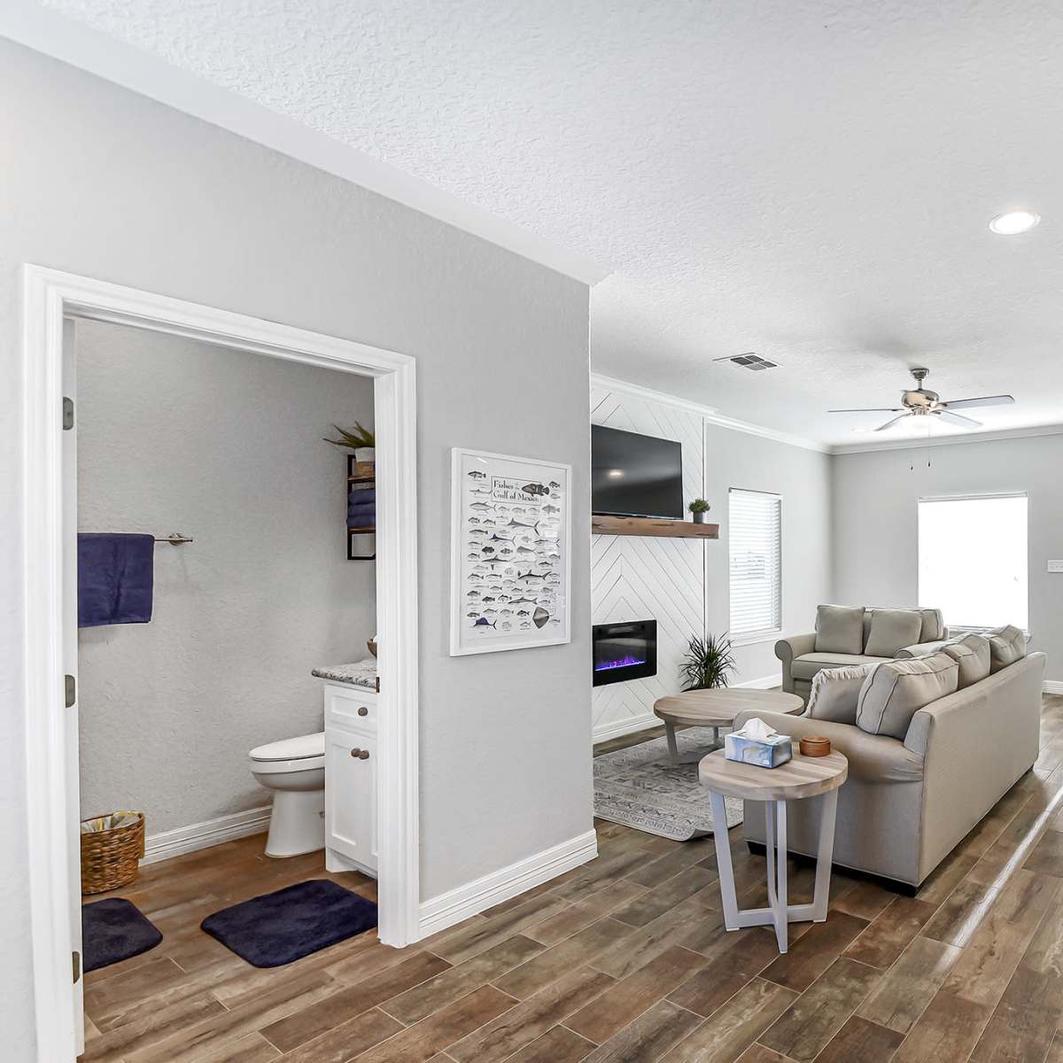 A modern living room connected to a dining area, featuring light-colored walls, wooden flooring, and a bathroom accessible from the living space.