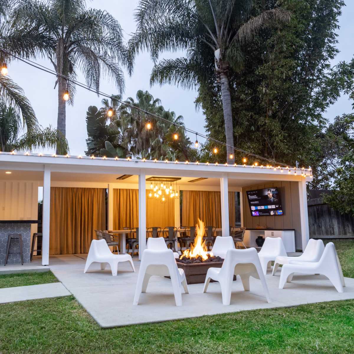 An outdoor patio area with seating around a fire pit, string lights overhead, and a television mounted on the wall.