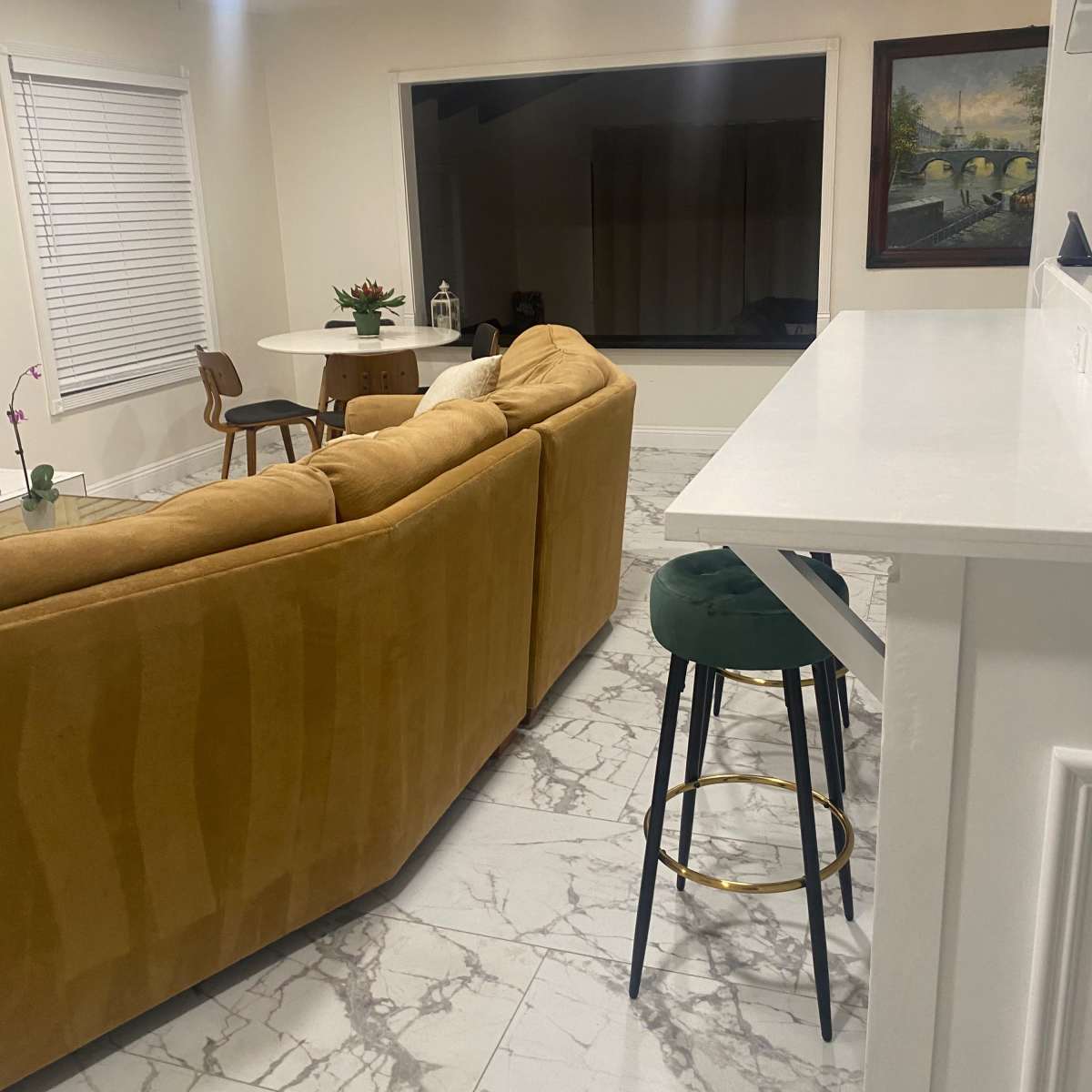 A living area with a yellow sofa, a round dining table, and a large window overlooking a darkened space.