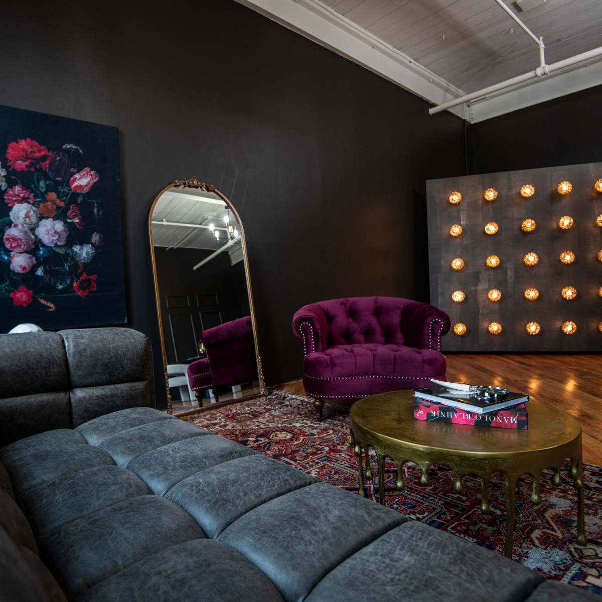 A stylish interior with a tufted purple armchair, a velvet grey sofa, a large mirror, and a lighted wall panel, complemented by floral artwork and a patterned rug.