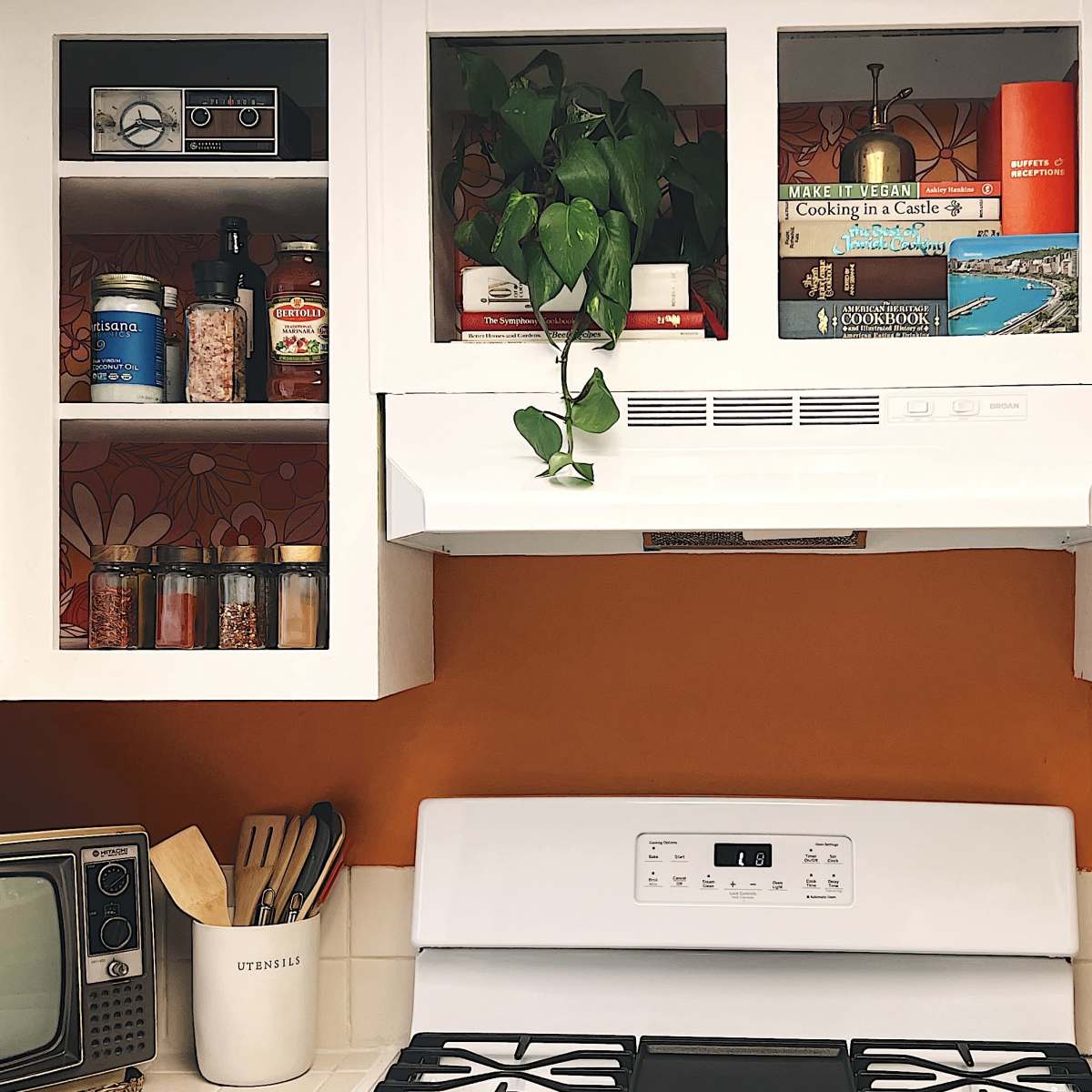 A kitchen scene features white cabinets with glass doors displaying jars and books, a white stove beneath a hood, and a vintage television on the counter.
