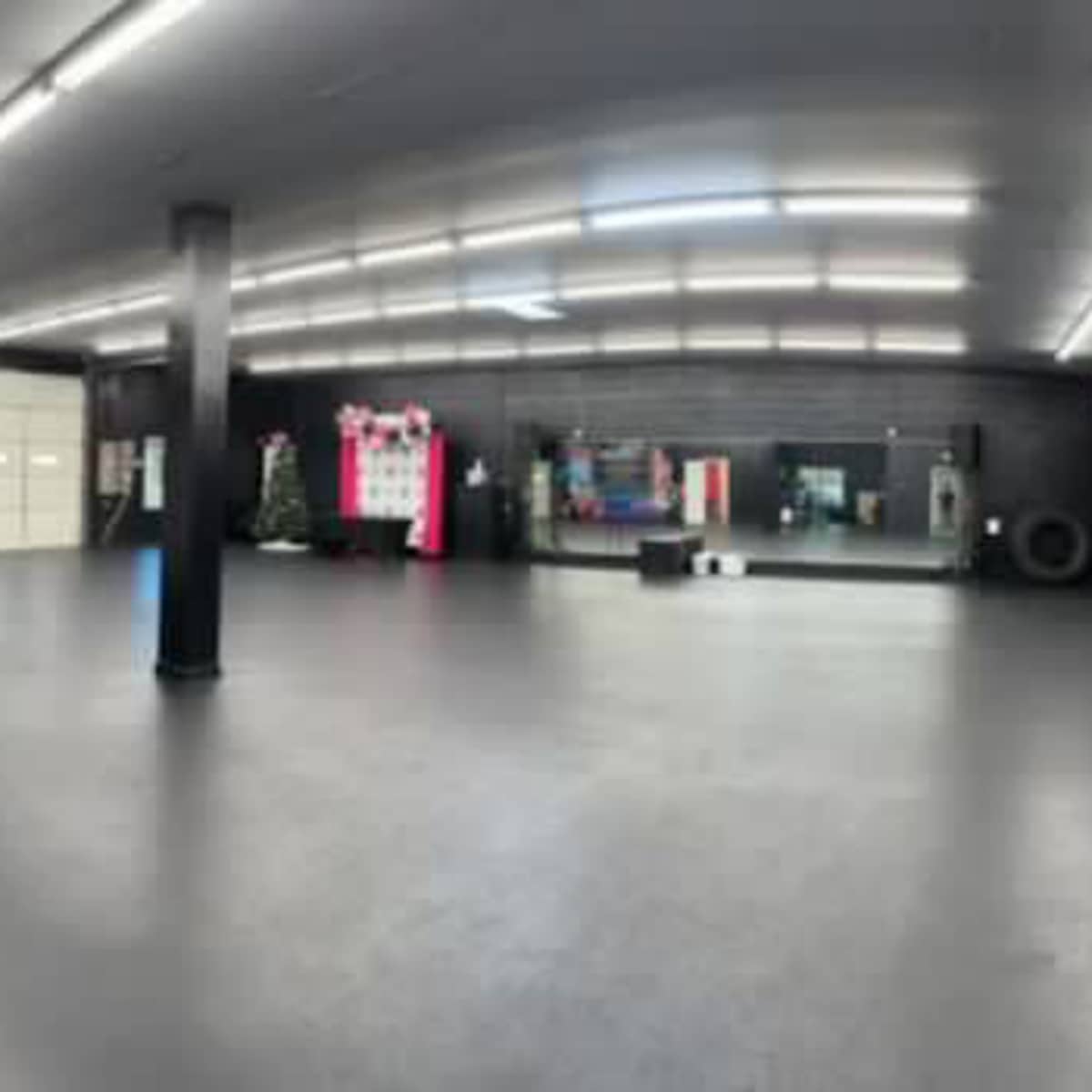 A spacious gym interior with dark walls, large rubber flooring, and various fitness equipment arranged along the sides.