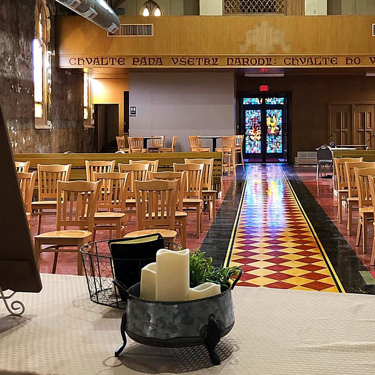 A spacious, empty hall with wooden chairs arranged in rows and a colorful stained glass window at the far end.