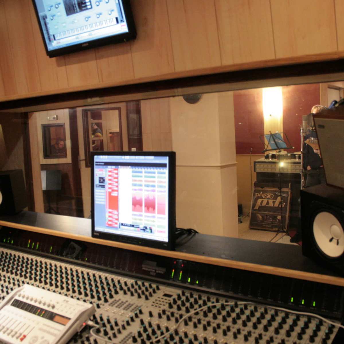 A recording studio control room features a large mixing console, computer monitor displaying audio tracks, and speakers, with a glimpse of the live room visible through a glass window.