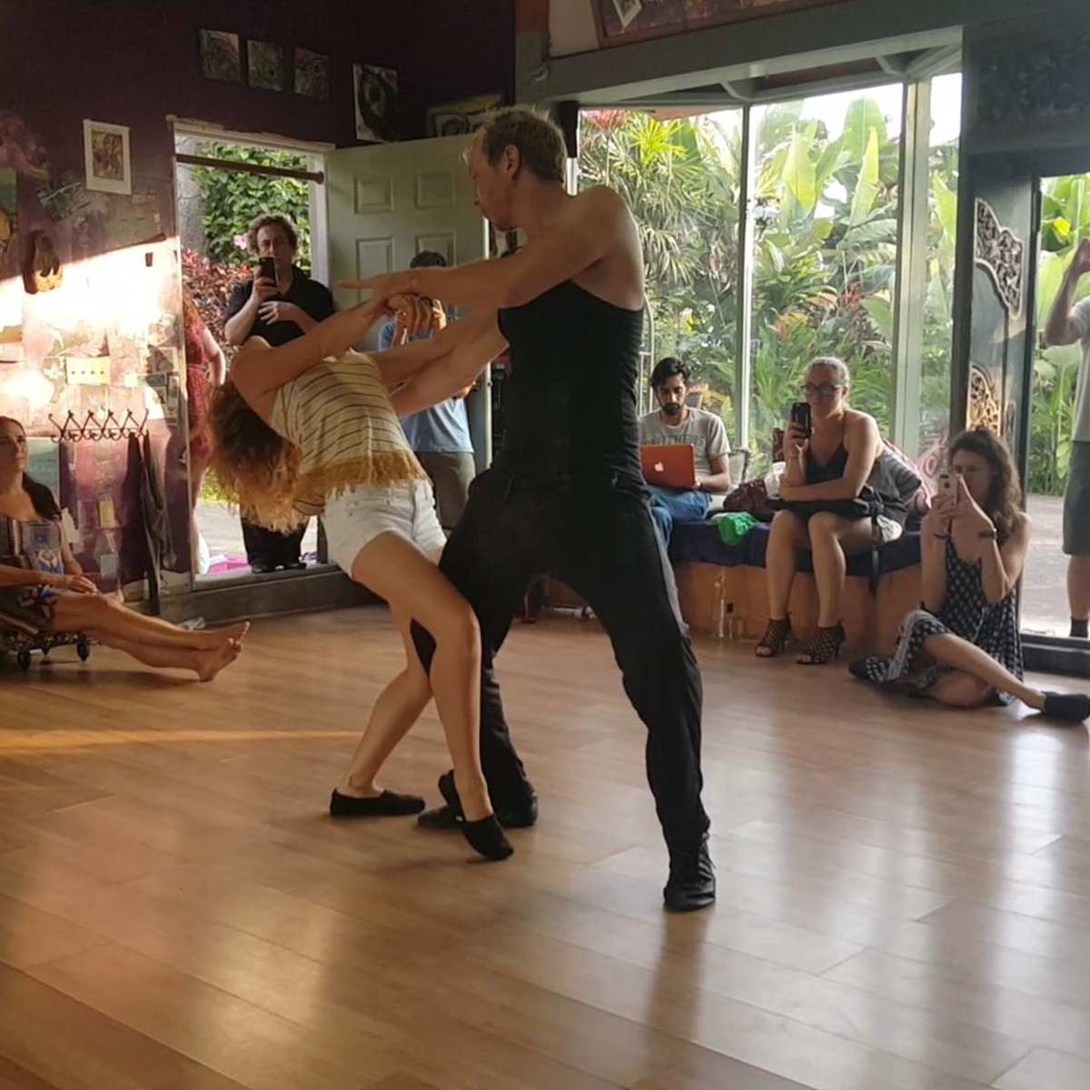 Two dancers perform a routine in a studio with spectators observing from various spots around the room.