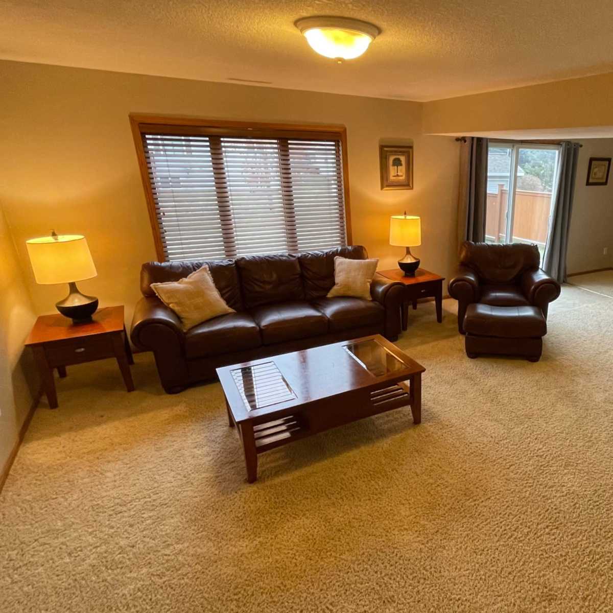 A living room with a seating area that includes a brown leather sofa, an armchair, two lamps on side tables, and a view of a sliding glass door leading to an outdoor space.