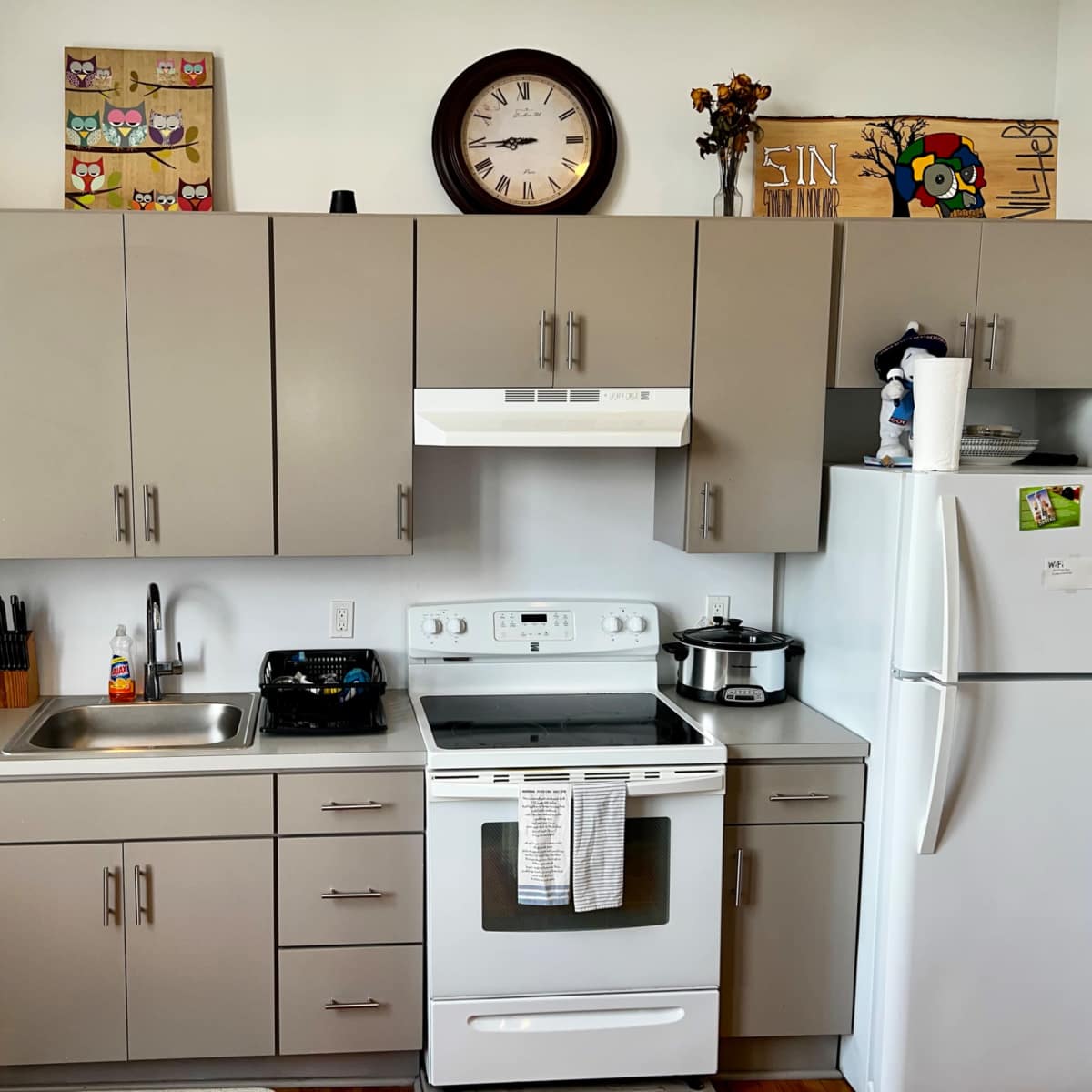 A modern kitchen with gray cabinetry, a stove, a microwave, and a refrigerator, along with a clock and decorative artwork on the walls.