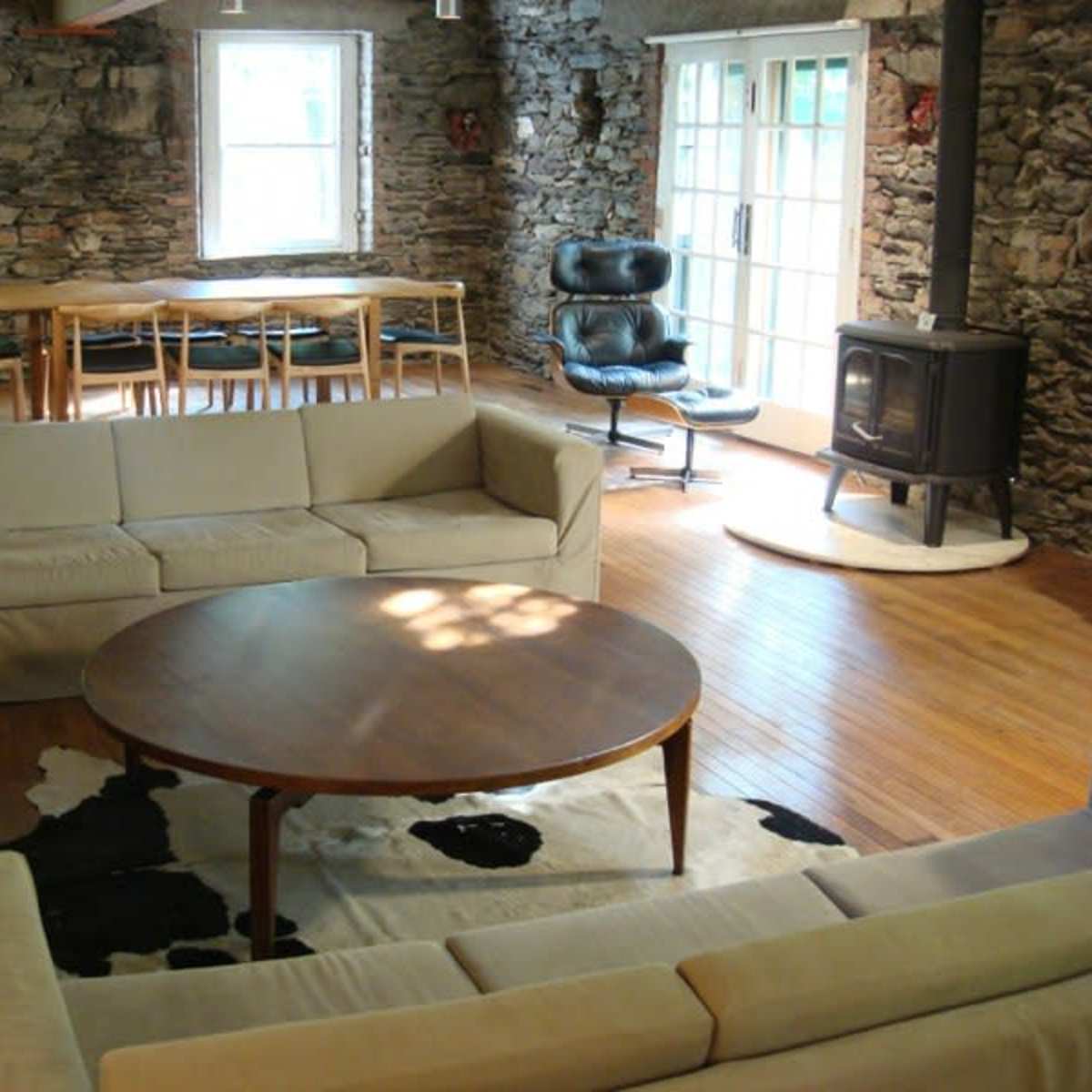 A spacious living room featuring stone walls, wooden floors, a circular table, and a cozy seating area with a sofa and an armchair near a wood stove.