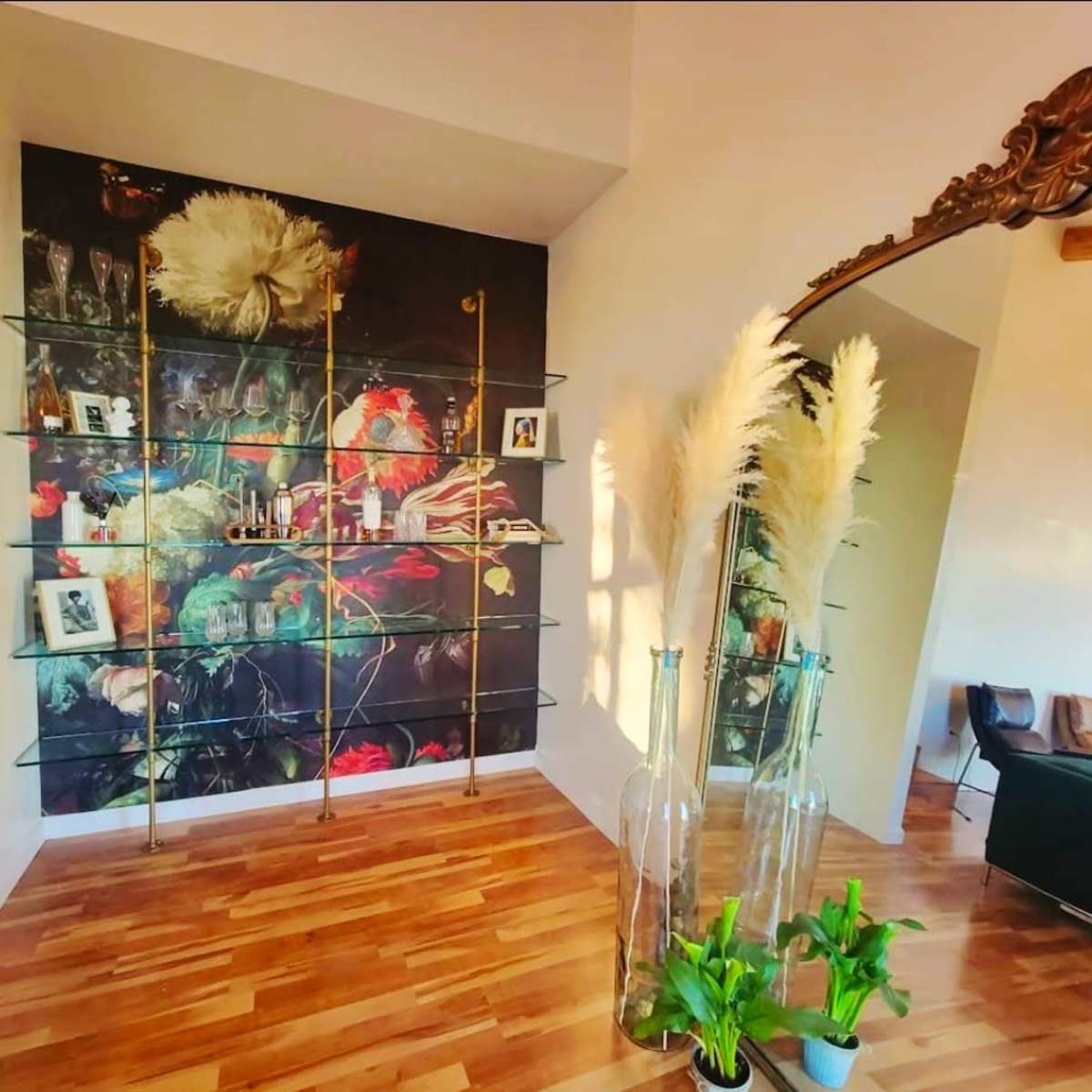 A modern interior featuring a glass shelf displaying decorative items and framed photos against a dark floral wall mural, complemented by tall vases and a green plant in the foreground.