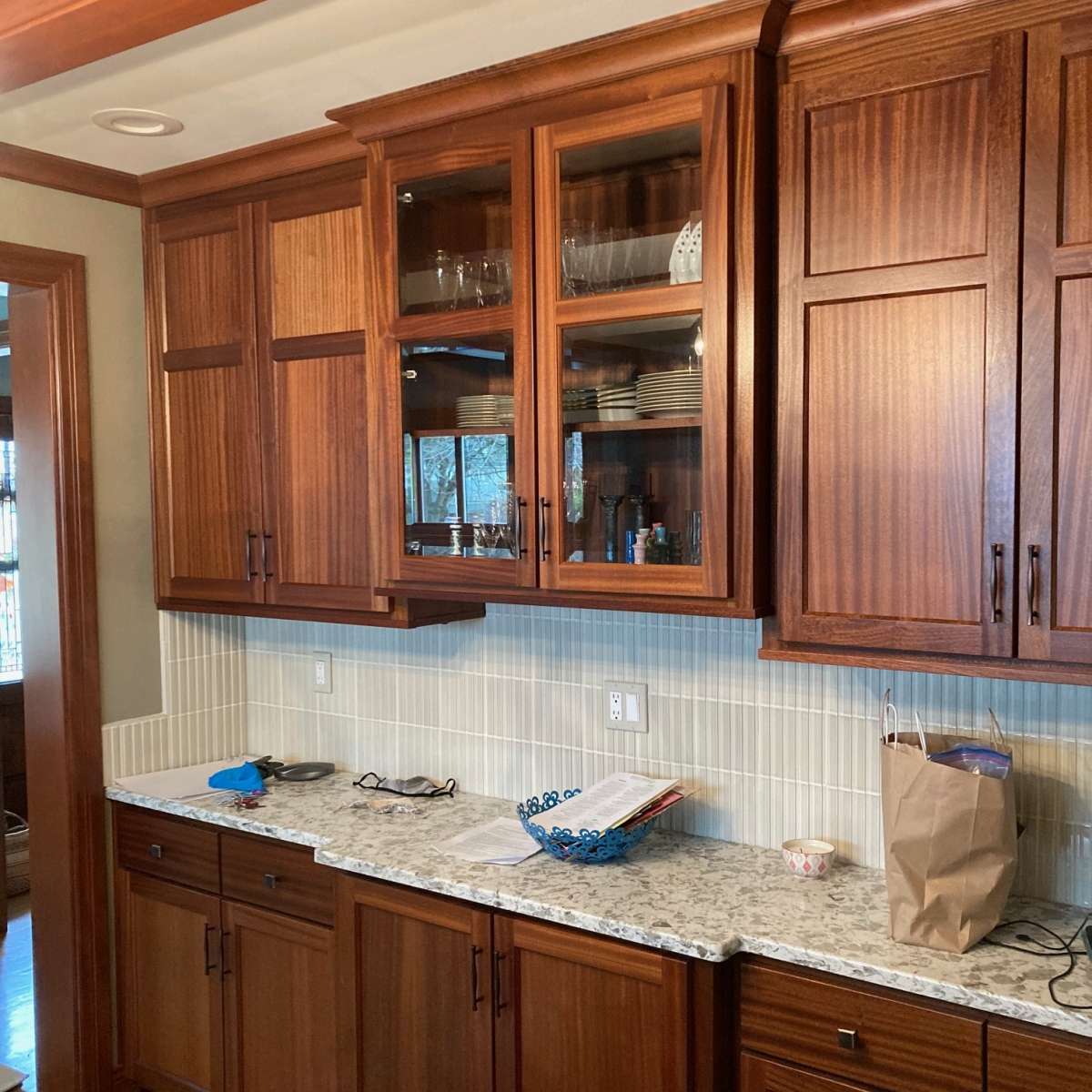 A kitchen with wooden cabinets, a granite countertop, and a tiled backsplash, featuring glassware and dishes displayed in an upper cabinet.
