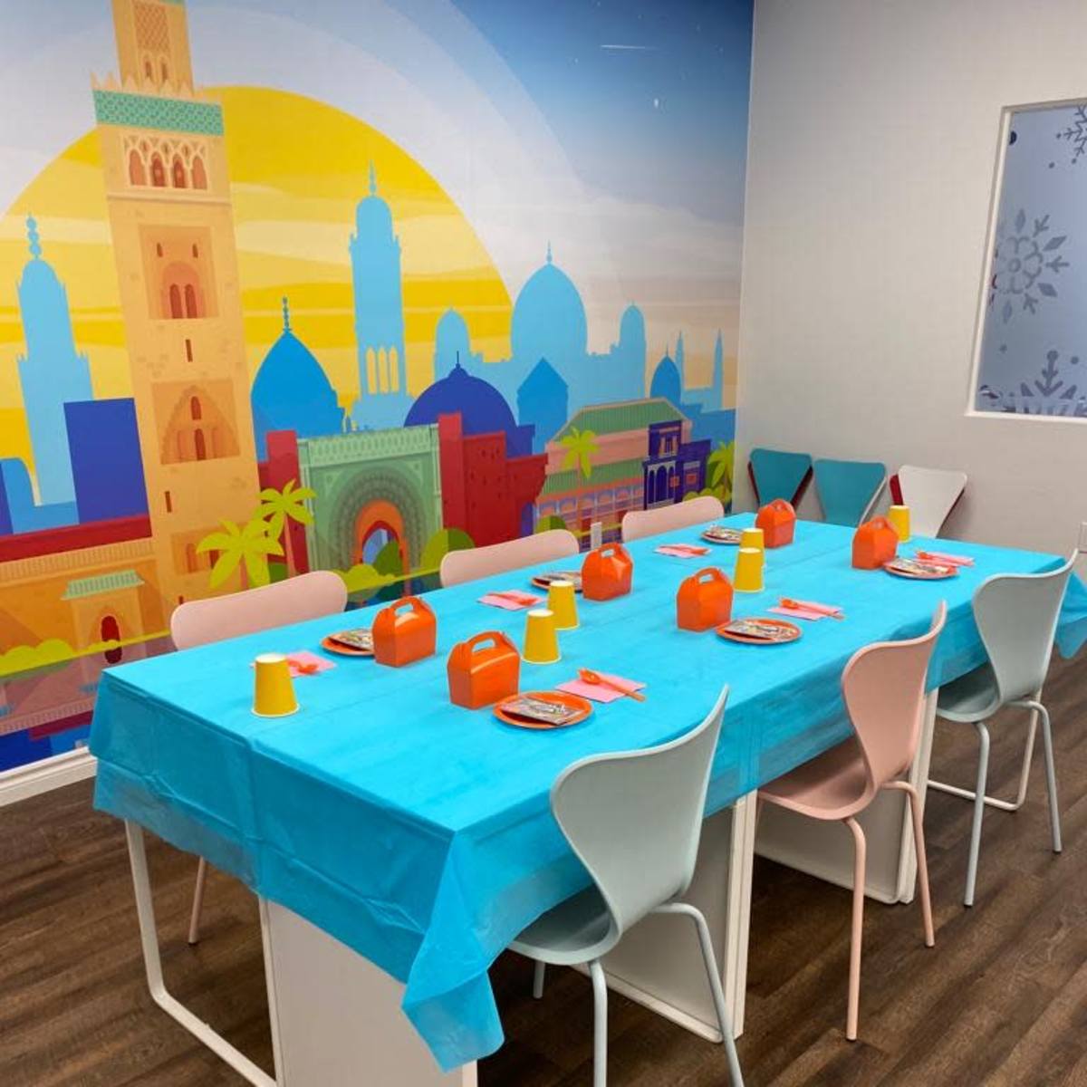 A large table is set with a blue tablecloth, colorful cups, and plates, surrounded by chairs in a room adorned with a vibrant mural.
