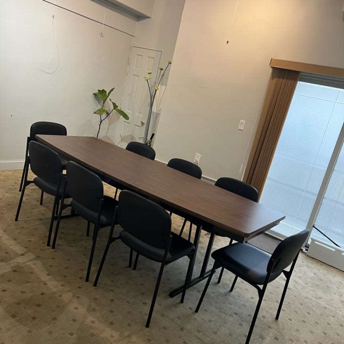 A rectangular wooden table surrounded by eight black chairs is positioned in a room with beige carpet and large windows.
