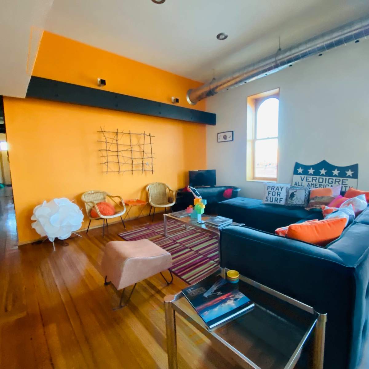 A modern living room with orange walls, wooden flooring, a black sectional sofa, and two rattan chairs, along with decorative elements like a TV and artwork.