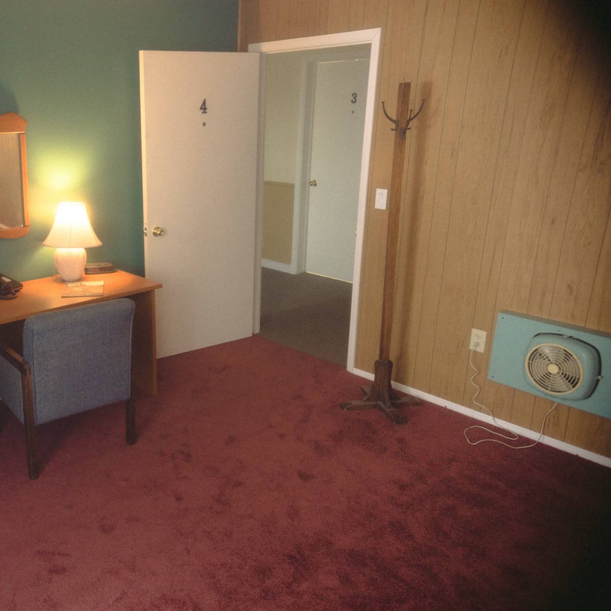 A hotel room features a desk with a chair, a lamp, a mirror, a mini fridge, and a television mounted on the wall, with an open door leading to another room.