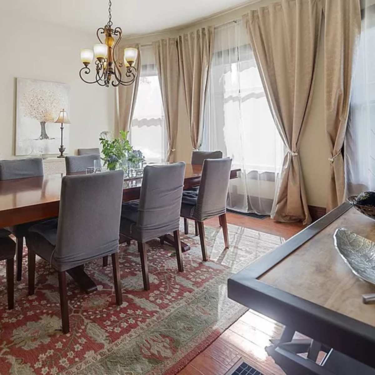 A dining room features a large wooden table surrounded by eight upholstered chairs, with large windows draped in curtains and a decorative chandelier overhead.