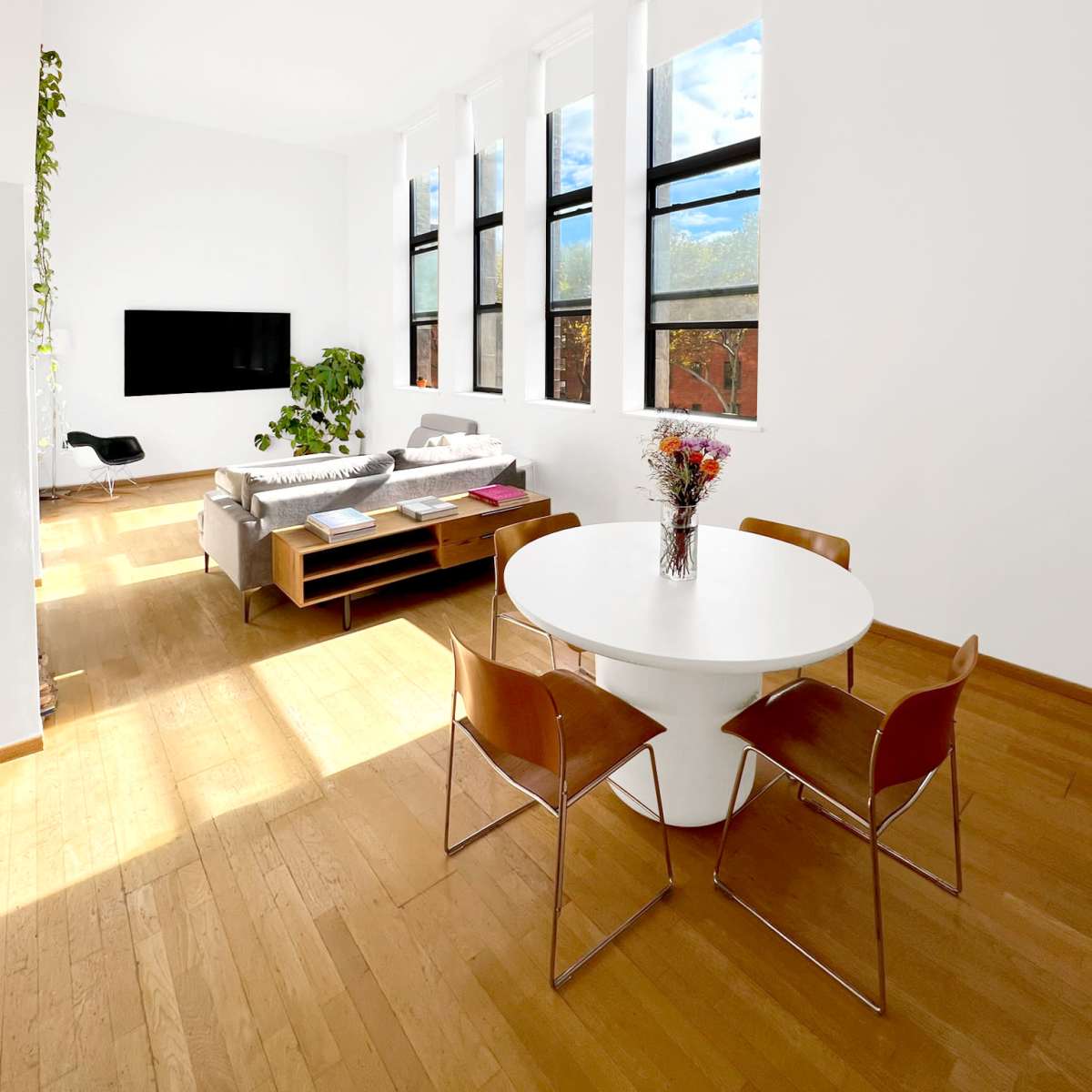 A modern living space with a round dining table and four chairs, a sofa, a television, and large windows letting in natural light.