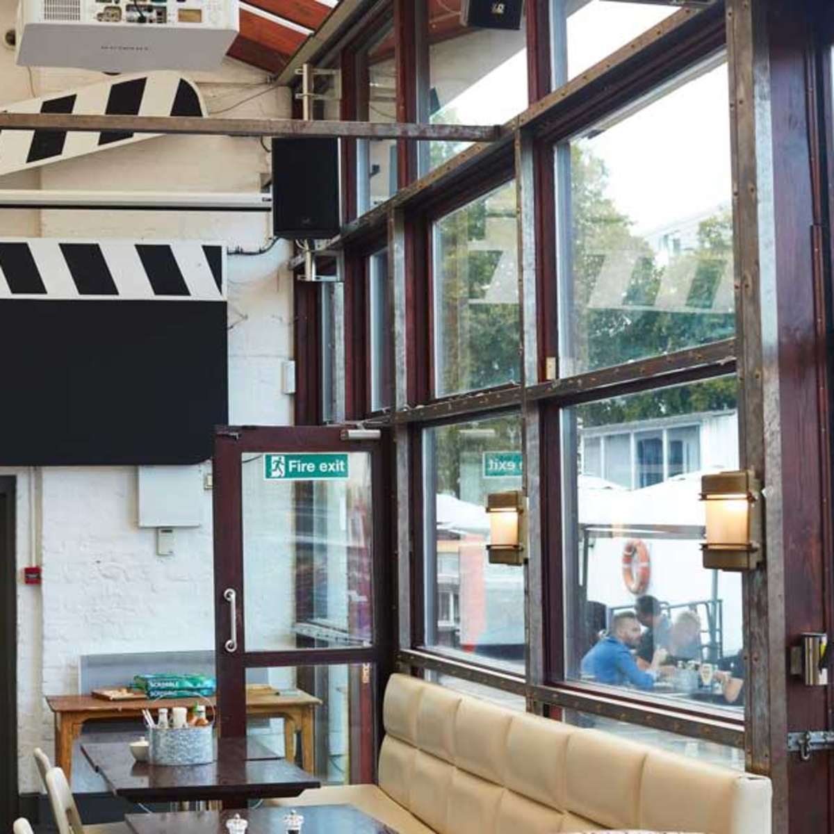 The interior of a restaurant features large windows, wooden furnishings, and a film clapperboard motif on the wall.
