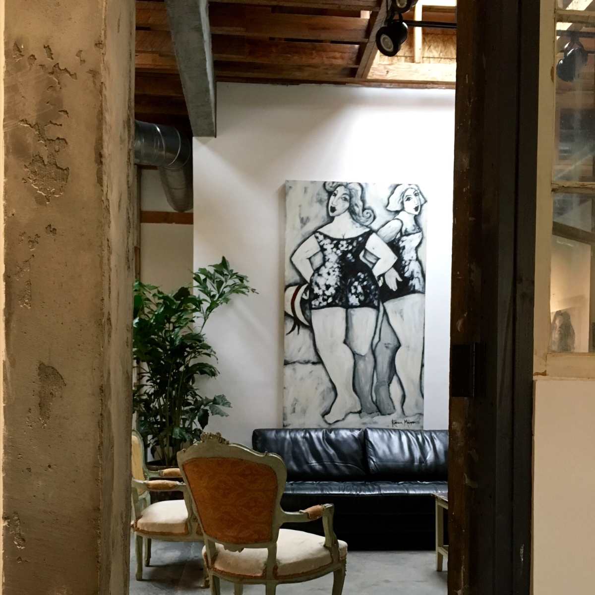 A hallway leads to a sitting area with a black leather couch, two chairs, a large artwork on the wall, and a potted plant.
