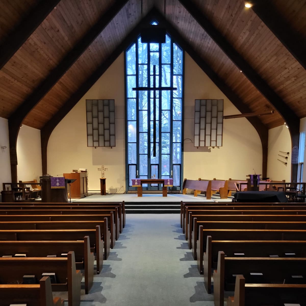 The interior of a church features wooden beams, rows of pews, and a large stained glass window at the front.