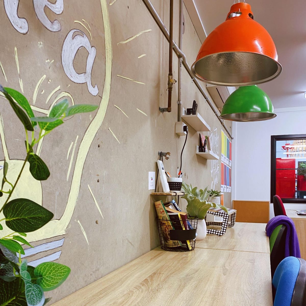 A modern workspace with a wooden table, colorful pendant lights hanging overhead, and a concrete wall featuring a light bulb drawing.