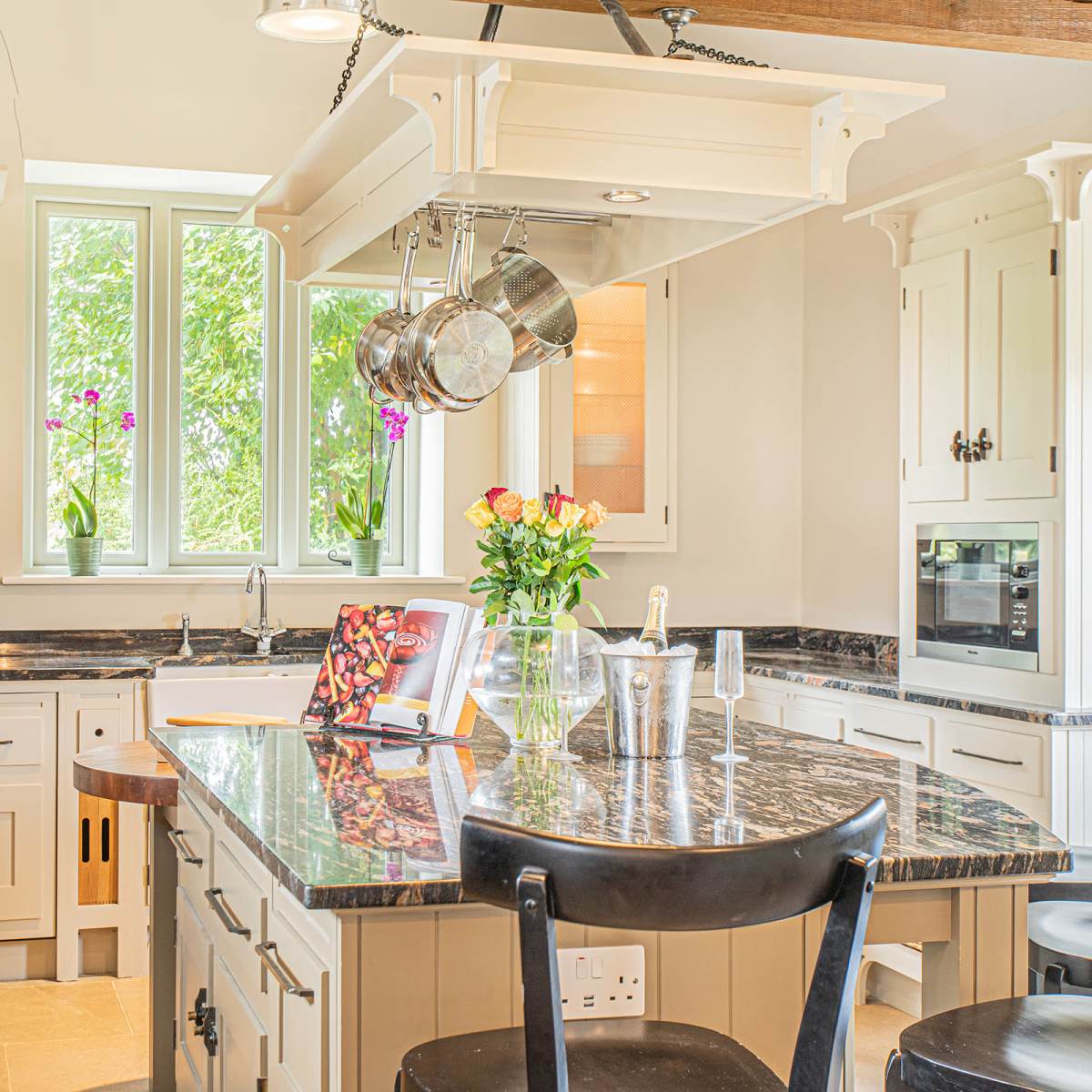 The kitchen features a central island with a marble countertop, surrounded by black chairs, and includes windows that provide natural light and view of greenery outside.