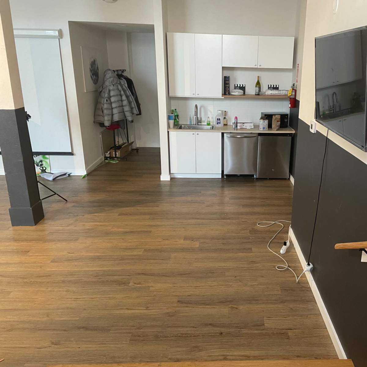 A spacious indoor area with a kitchen in the background, featuring a stainless steel refrigerator and white cabinetry, and a staircase leading downwards.