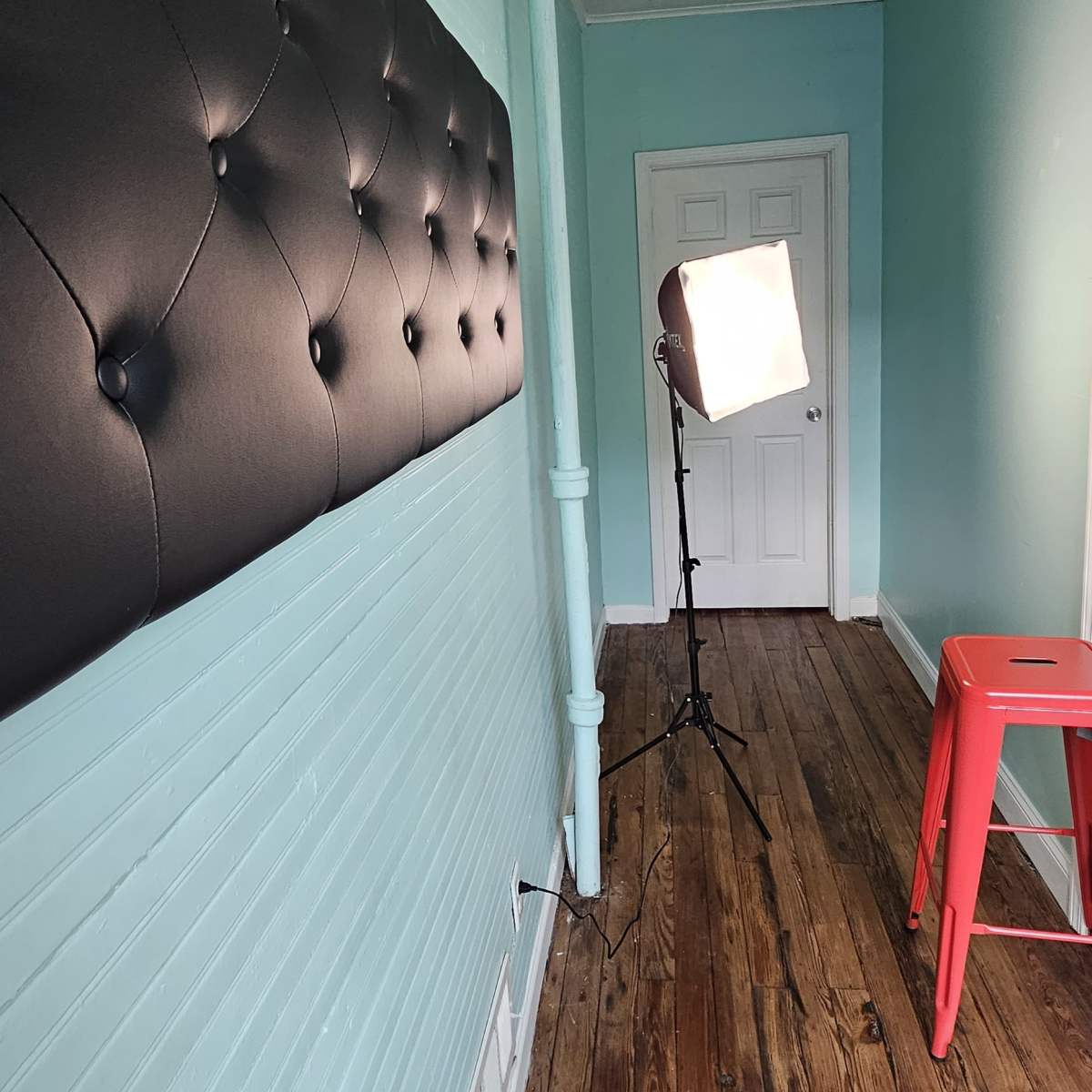 A narrow hallway with a light blue wall, a black tufted panel on one side, hardwood flooring, and a red stool near a photography light setup.