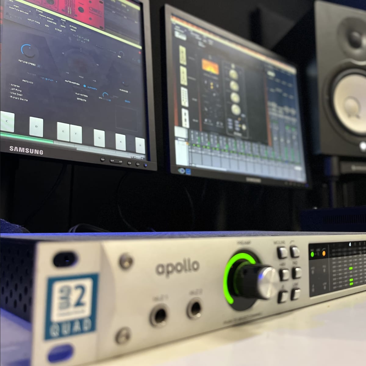 An Apollo audio interface positioned in front of two computer monitors displaying digital audio workstation software, with a studio monitor speaker in the background.