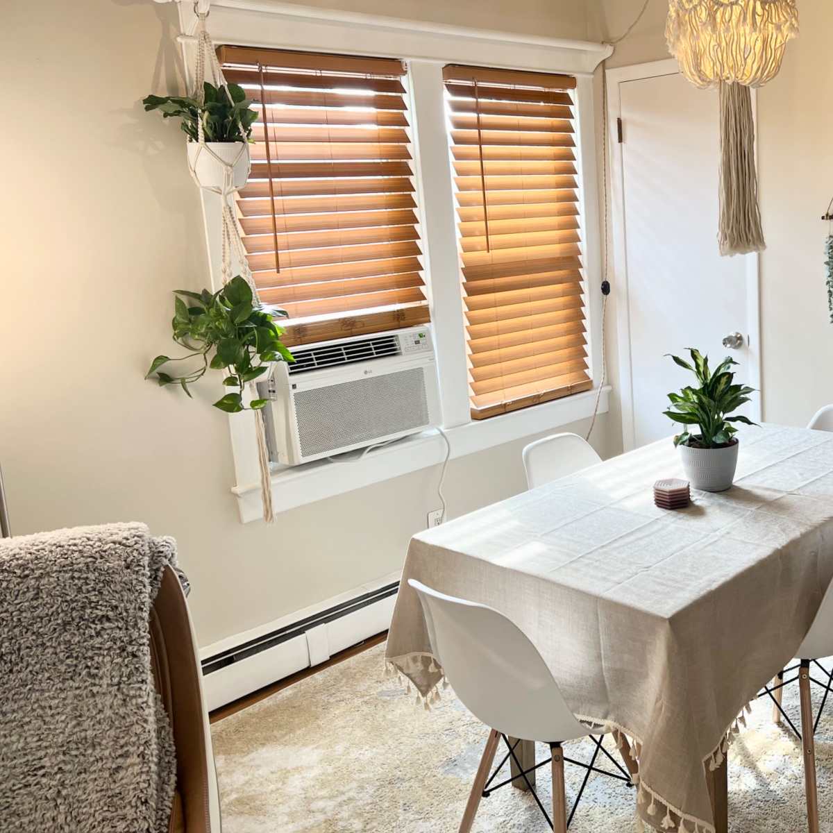 A dining area features a table with a cream-colored tablecloth, surrounded by white chairs, and is adorned with hanging plants and window blinds.
