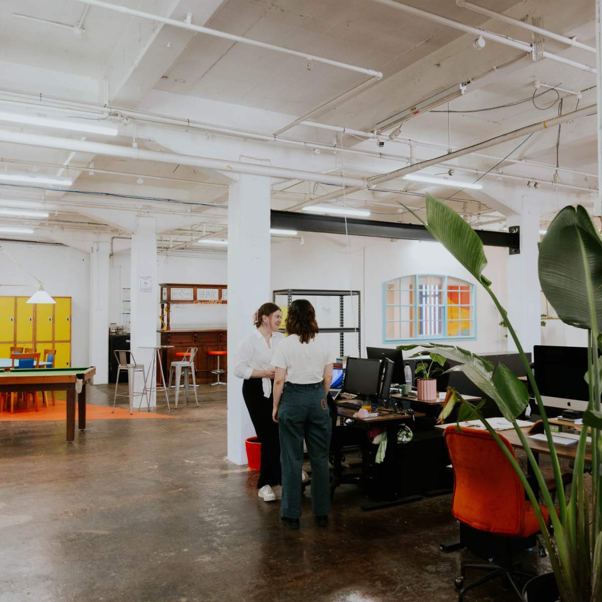 Two people are standing in an open office space with computers, plants, and colorful decor.