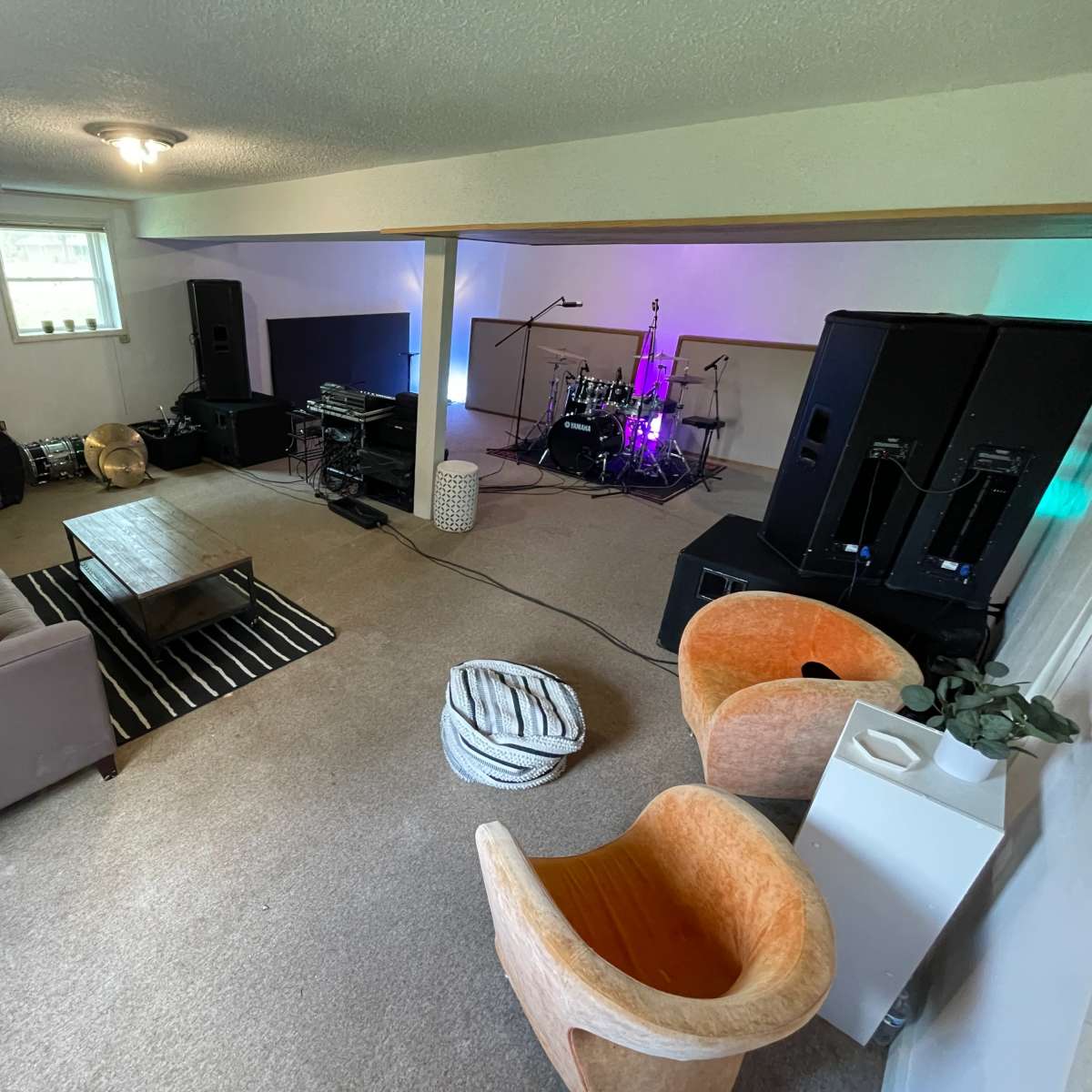 A spacious basement area with a music setup featuring a drum kit, amplifiers, and seating arrangements including a sofa and orange chairs.