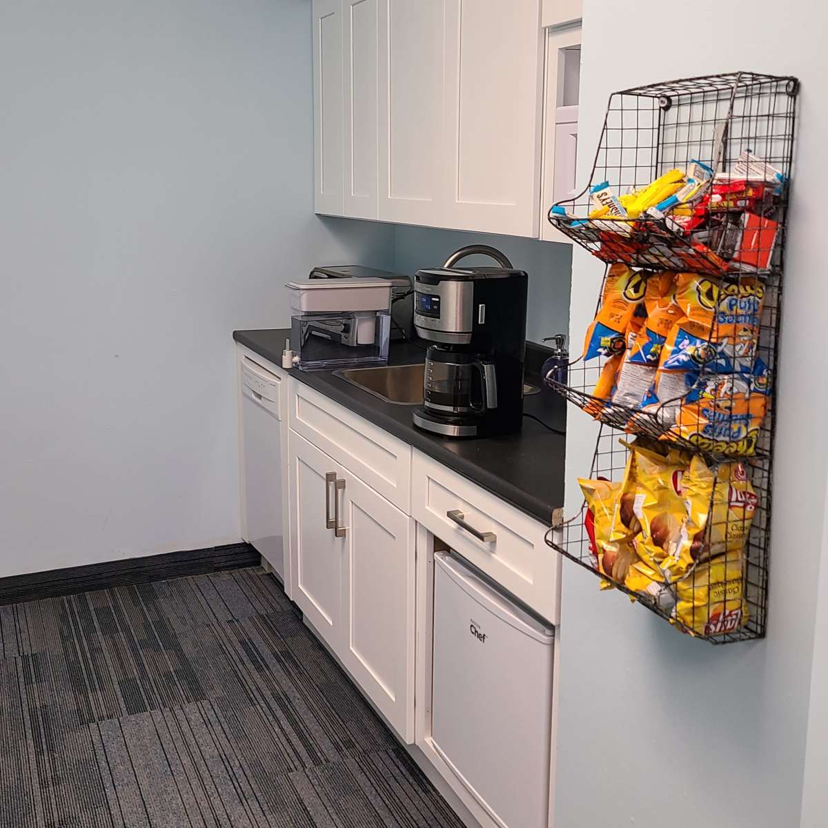 A kitchen area with a dark countertop, a coffee maker, a sink, and a wall-mounted wire rack filled with snack bags.