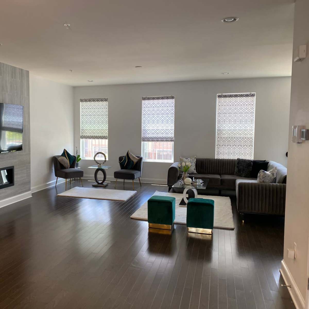 A modern living room with a large flat-screen TV mounted on a gray wall, a fireplace beneath it, and a sectional sofa surrounded by two armchairs and glass coffee tables.