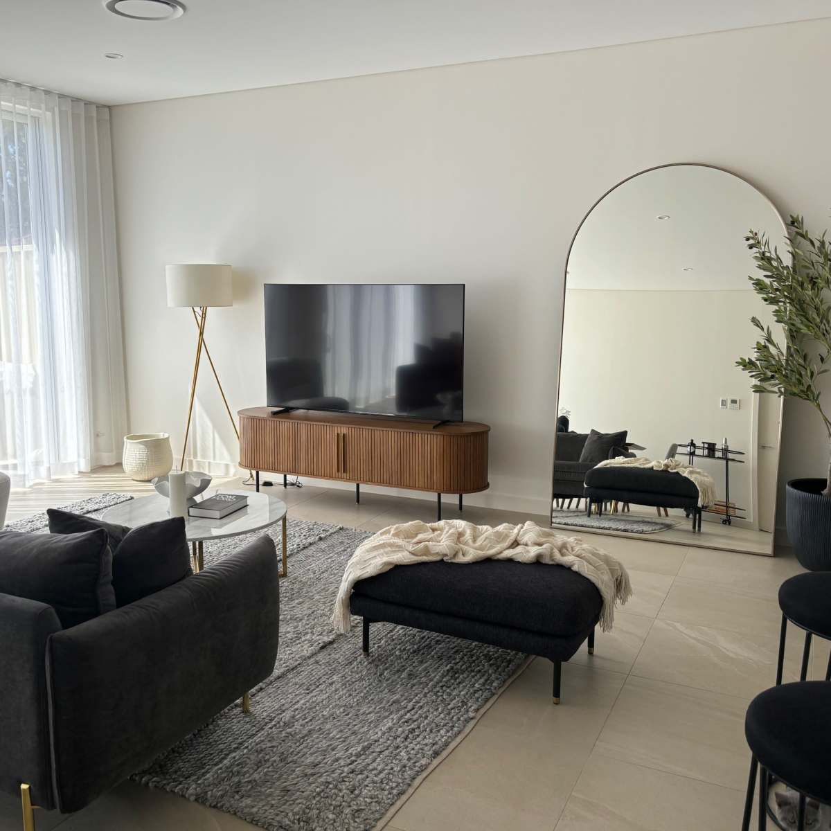 The living room features a dark sofa, a wooden TV cabinet, a large mirror, and a contemporary lamp, with light streaming through sheer curtains.