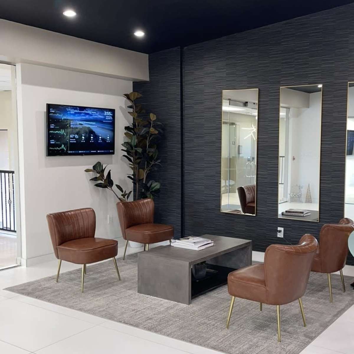 A modern waiting area featuring four brown leather chairs arranged around a metal table on a light gray carpet, with a television mounted on the wall and two mirrors flanking it.