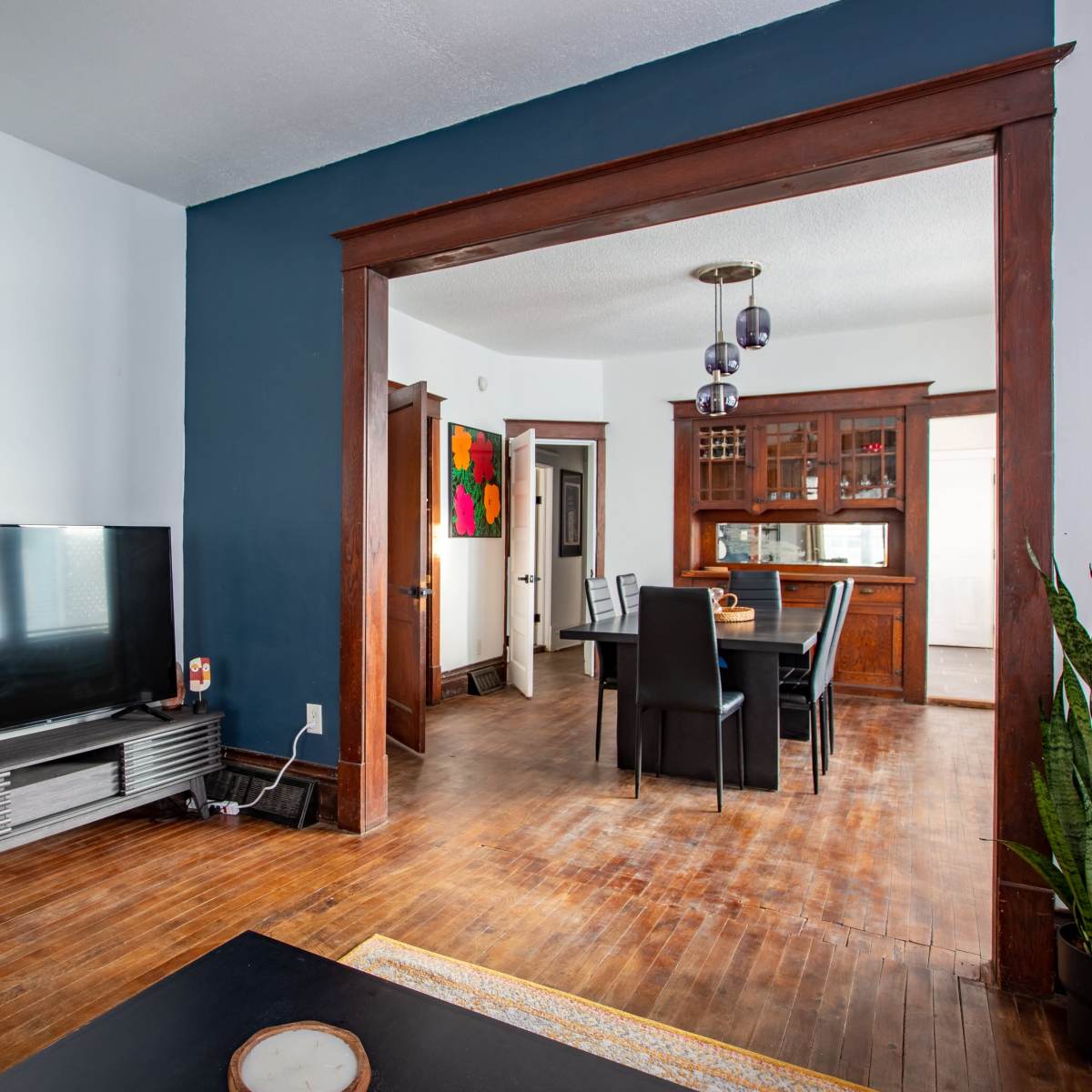 A living room with a television on a stand, hardwood floors, and a dining area visible in the background with a table and chairs.