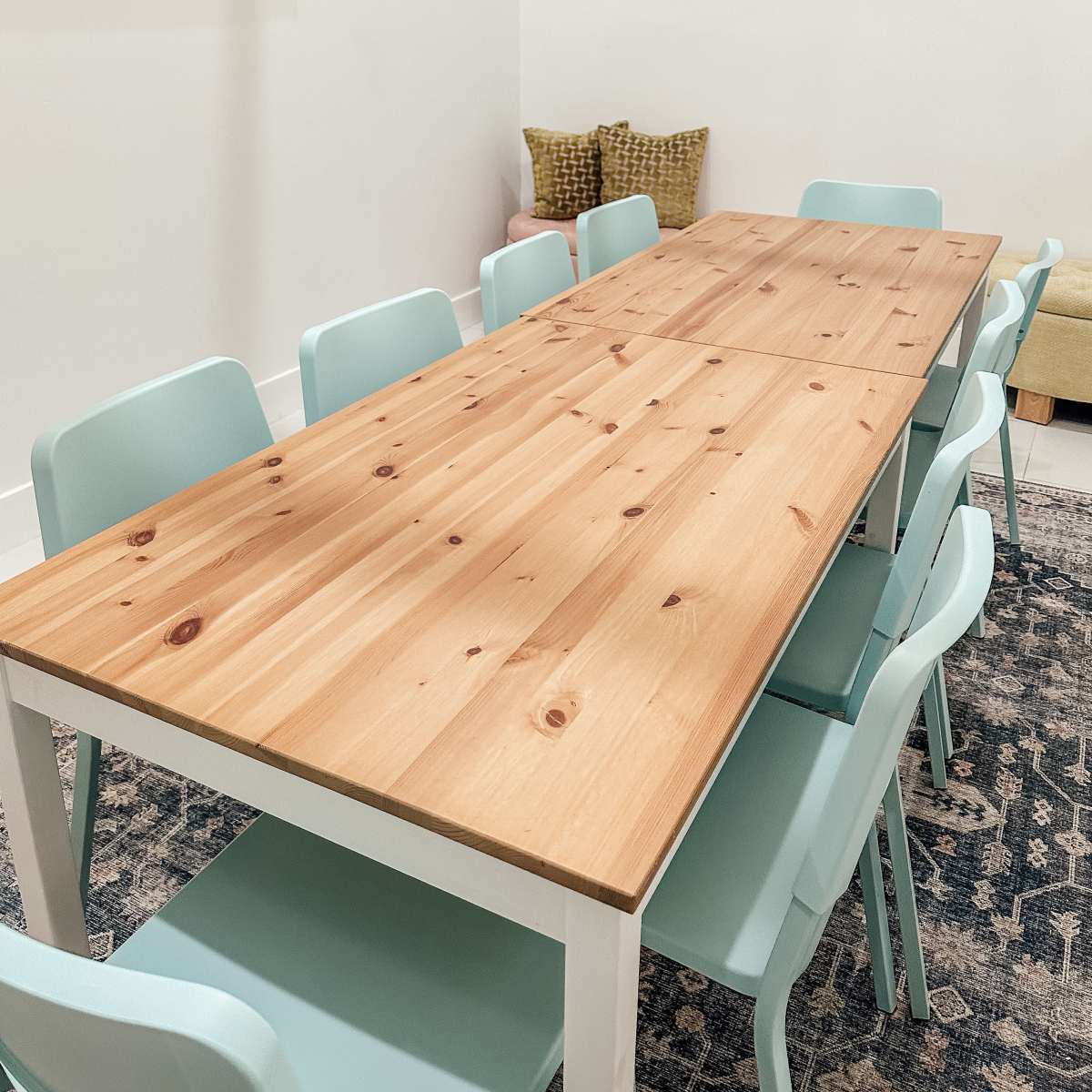 A long wooden table with a light finish is surrounded by several mint-colored chairs in a bright room with minimal decor.
