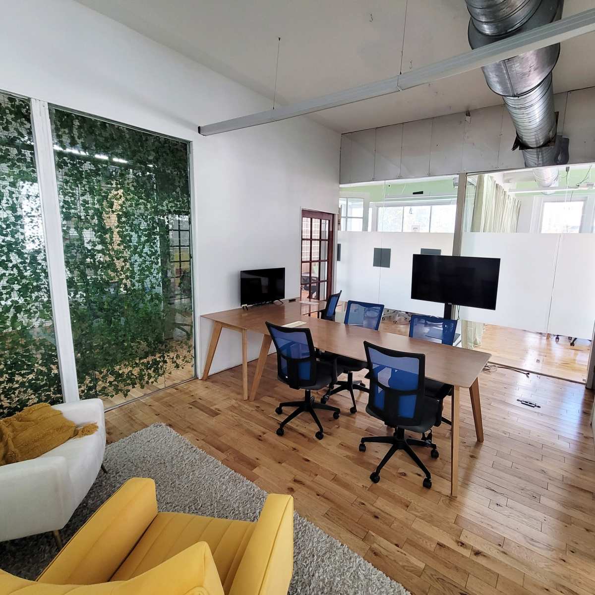 A modern office space featuring a conference table with four chairs, two large screens, and a seating area with yellow and white furniture, surrounded by glass walls adorned with greenery.