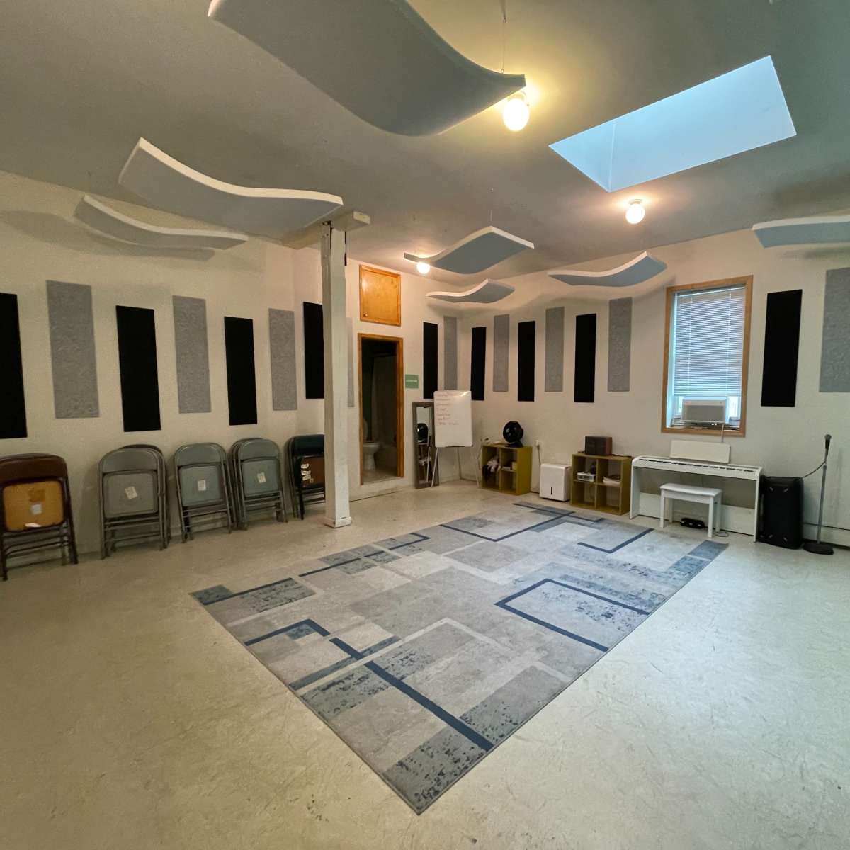 A spacious room with a light-colored floor, various sound-absorbing panels on the walls, a patterned rug in the center, and a set of folding chairs arranged on one side.