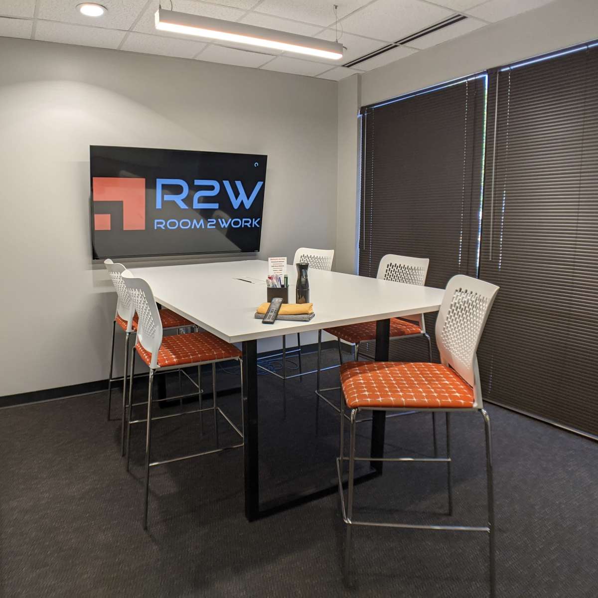 A modern meeting room with a rectangular table surrounded by six high-backed stools and a screen displaying the "R2W ROOM2WORK" logo on the wall.