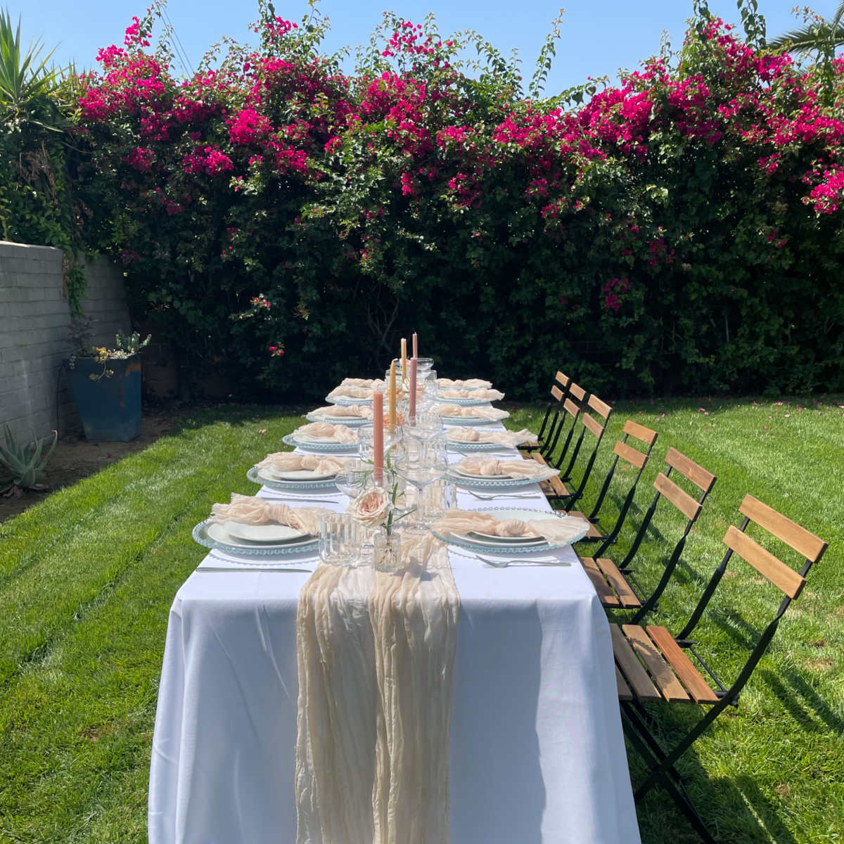 A long table is set for dinner with white cloth, plates, glasses, and pink candles, surrounded by green grass and flowering bushes.