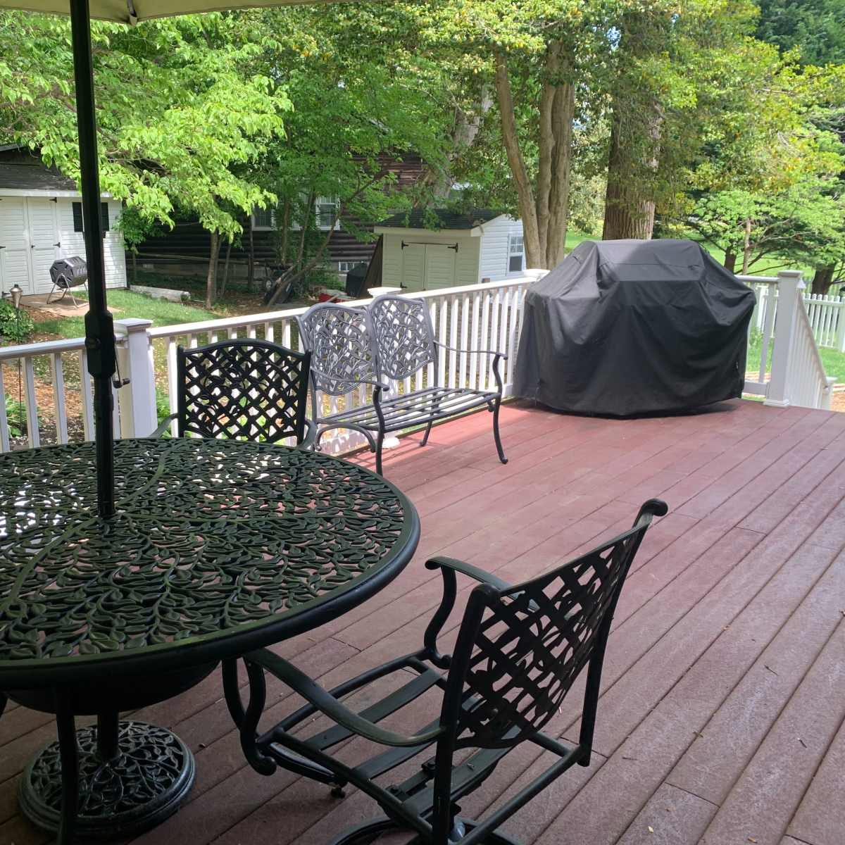 A patio area features a round metal table with chairs, an umbrella, and a grill, surrounded by lush greenery.