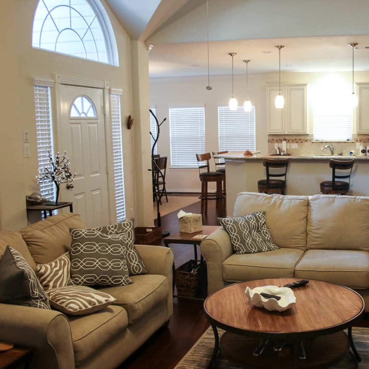 A spacious living room with a beige sofa, a coffee table, and an open layout leading to a kitchen area with white cabinetry and light fixtures.