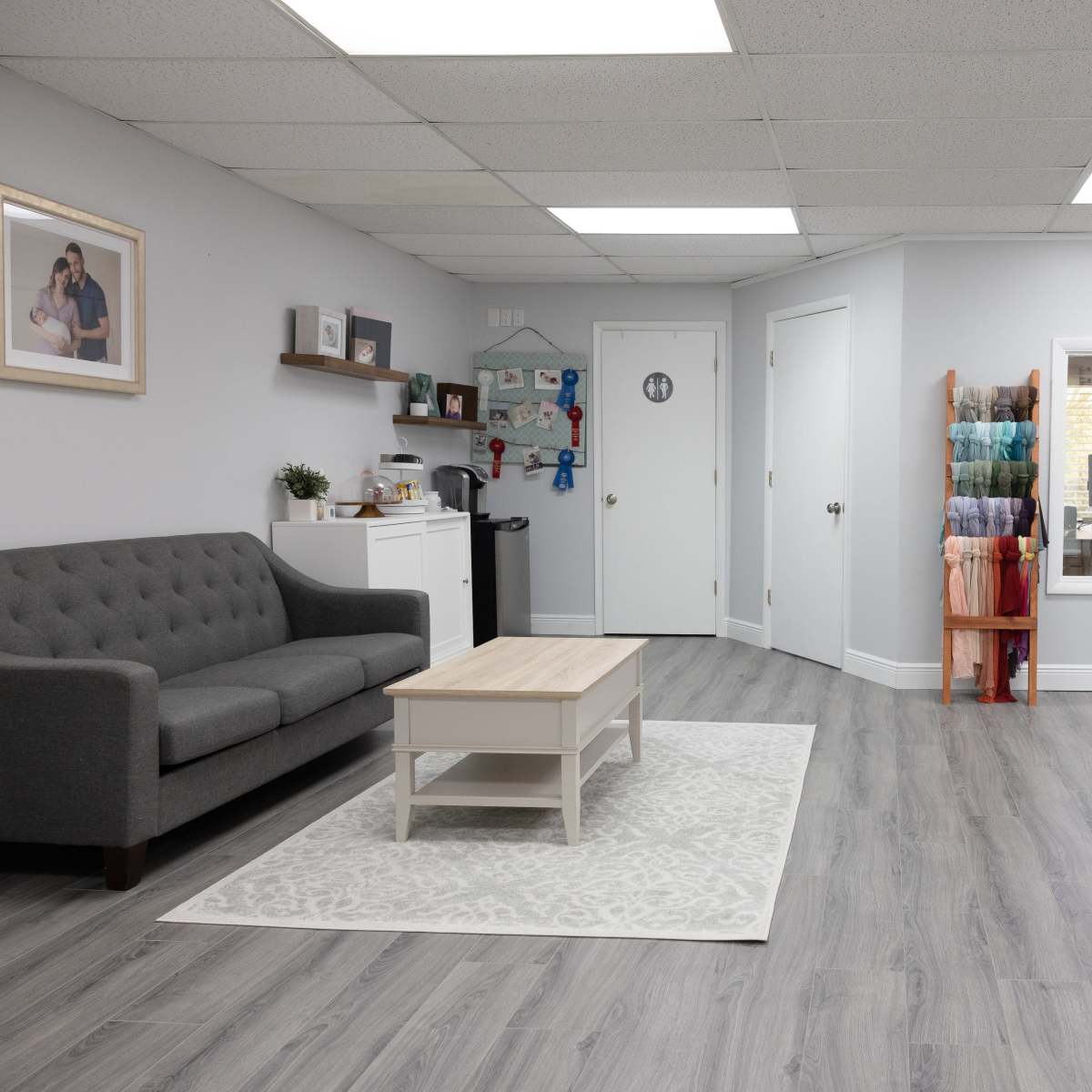 The room features a gray sofa and a wooden coffee table on a light rug, with colorful fabric rolls displayed on racks against the walls.