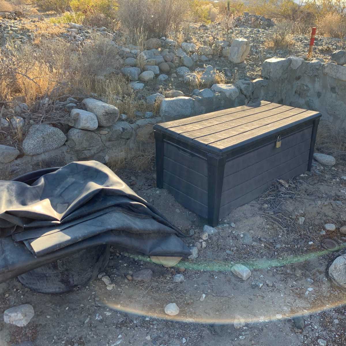 A wooden storage box is placed on rocky ground surrounded by sparse vegetation and stone structures.