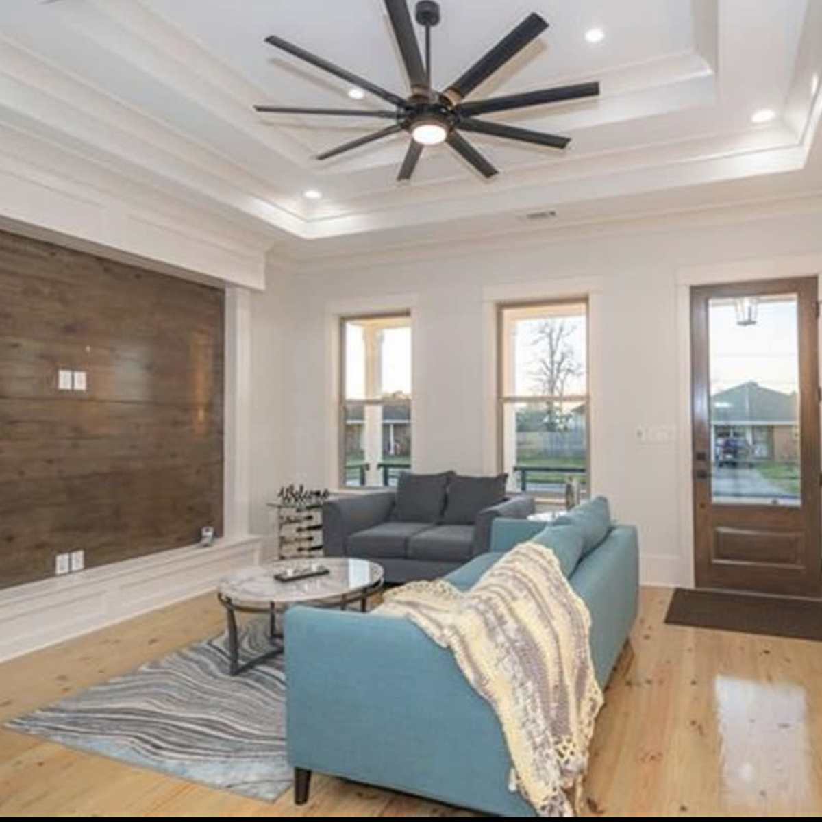 A modern living room with a blue sofa, a coffee table, and a large ceiling fan, complemented by natural light from the windows and a wooden accent wall.