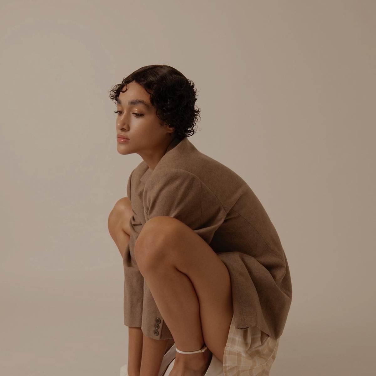 A person with short, curly hair kneels in a squatting position while wearing a tailored brown blazer and white high-heeled shoes.